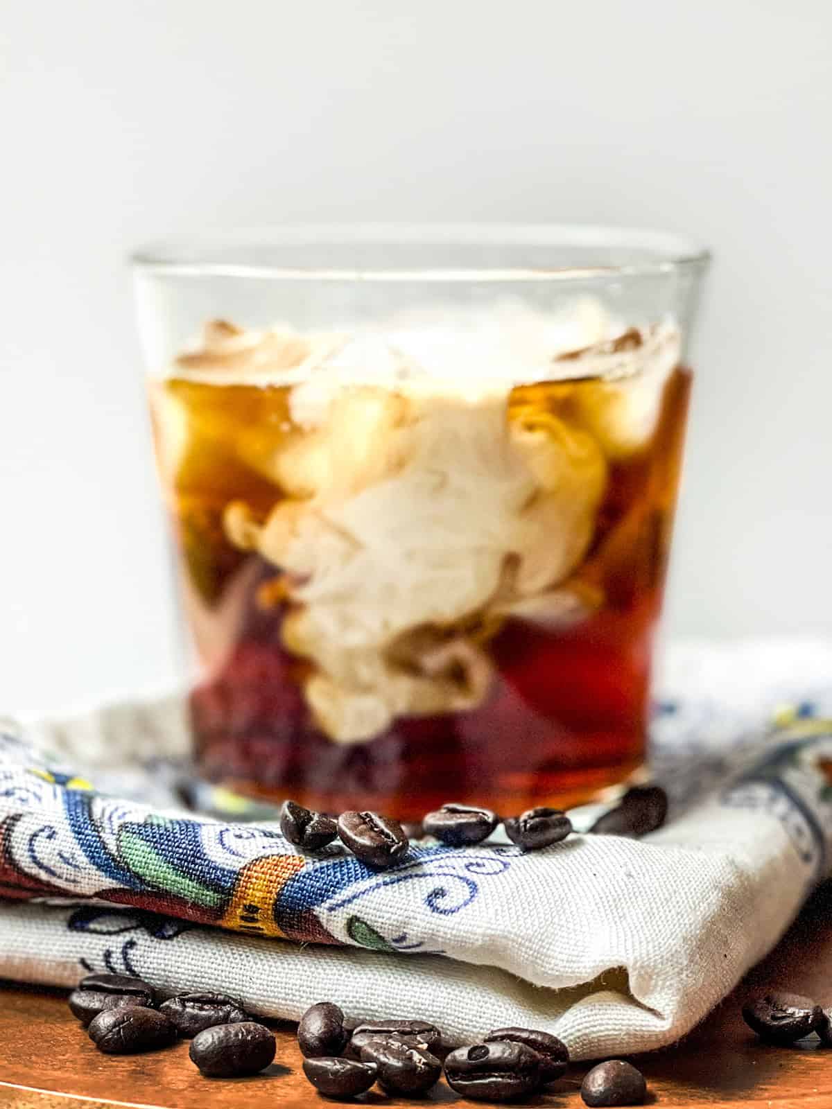 A glass of iced vanilla latte with cream swirling inside, placed on a patterned cloth, with coffee beans scattered in the foreground.