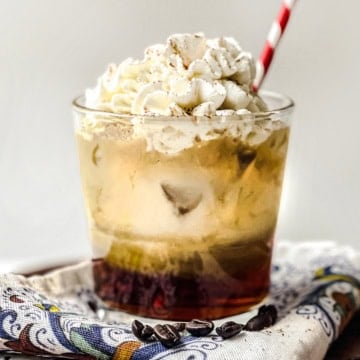 A glass of iced vanilla latte topped with whipped cream and a sprinkle of cinnamon, served with a red and white straw, placed on a patterned napkin with coffee beans nearby.