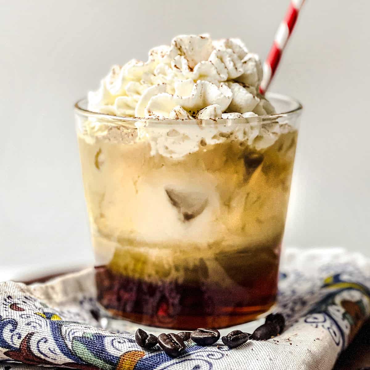A glass of iced vanilla latte topped with whipped cream and a sprinkle of cinnamon, served with a red and white straw, placed on a patterned napkin with coffee beans nearby.