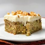 A slice of moist zucchini cake with white frosting and chopped walnuts on top, served on a white plate.
