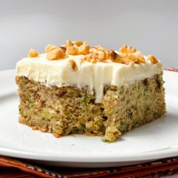 A slice of moist zucchini cake with white frosting and chopped walnuts on top, served on a white plate.