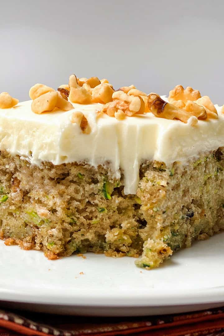 A slice of moist zucchini cake with white frosting and chopped walnuts on top, served on a white plate.