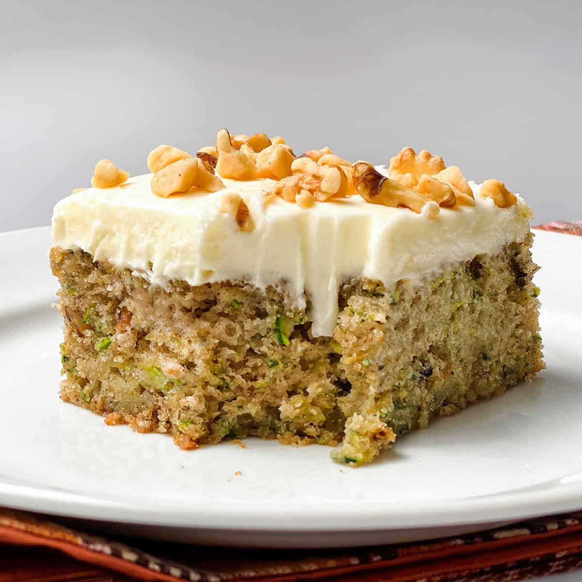 A slice of moist zucchini cake with white frosting and chopped walnuts on top, served on a white plate.