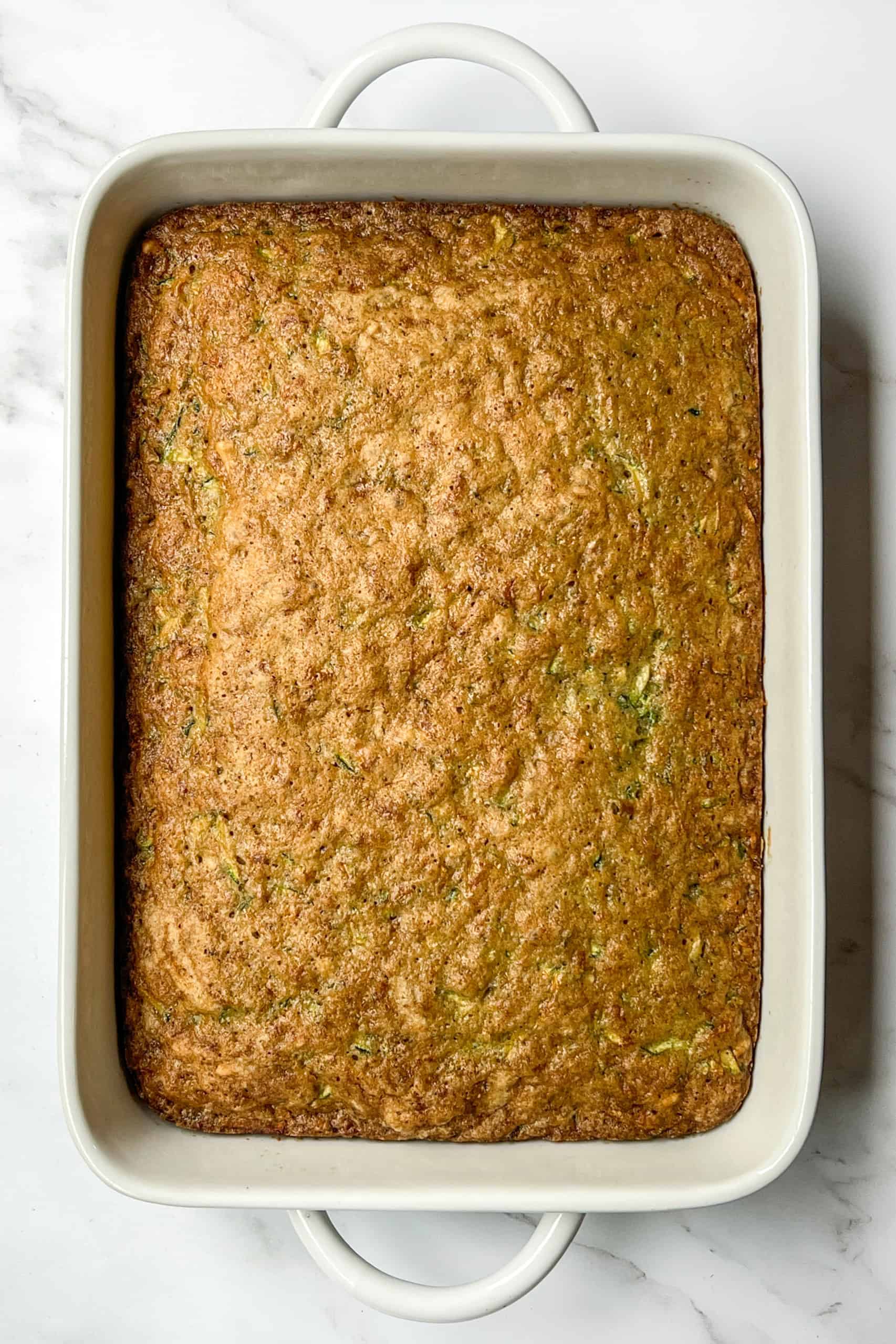 A rectangular baked zucchini cake rests in a white ceramic dish on a white marble surface.