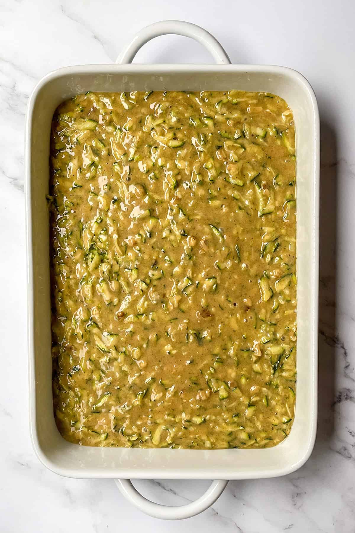 A white baking dish filled with raw zucchini cake batter sits on a marble surface, ready to be baked.