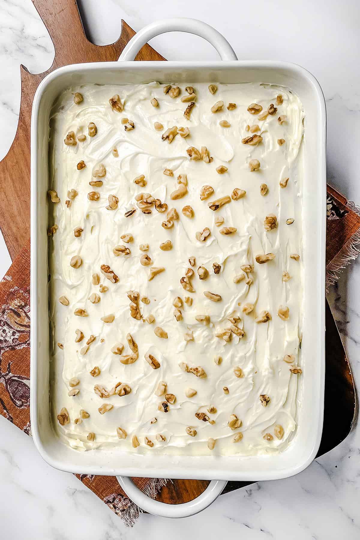 A rectangular baking dish with a zucchini cake topped with cream cheese frosting and scattered chopped walnuts, placed on a wooden board and patterned cloth.