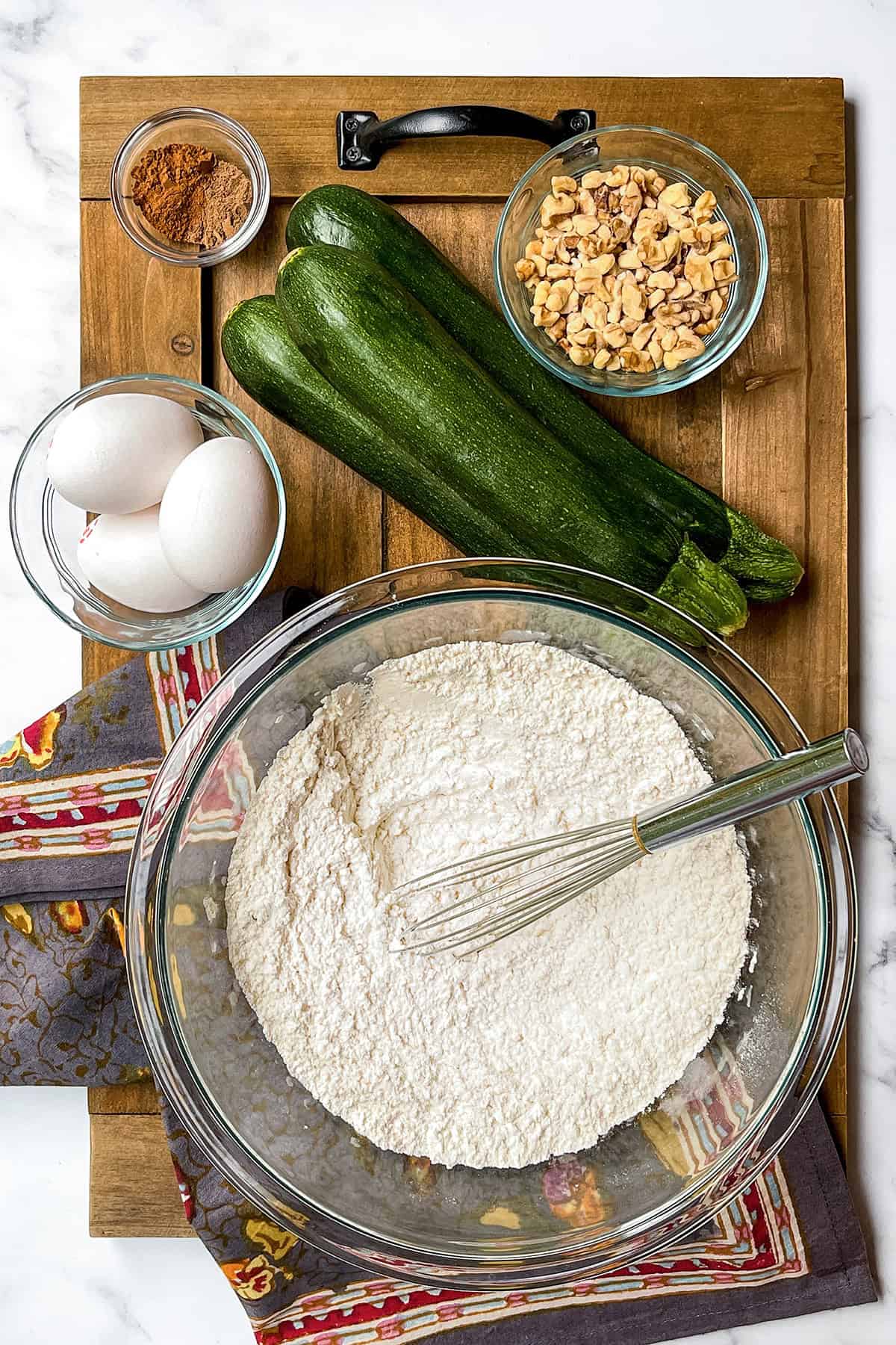 A glass bowl with flour and a whisk, two zucchinis, three eggs, chopped nuts, and a small bowl of cinnamon on a wooden board, all perfectly arranged and ready for baking a delicious zucchini cake.