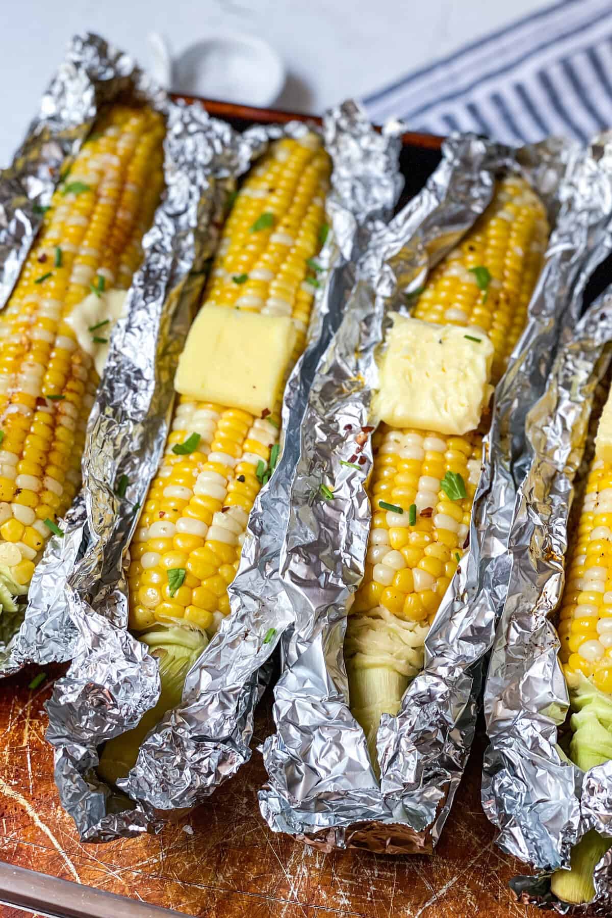 Four ears of grilled corn on the cob in aluminum foil, topped with butter and herbs, placed on a baking sheet.