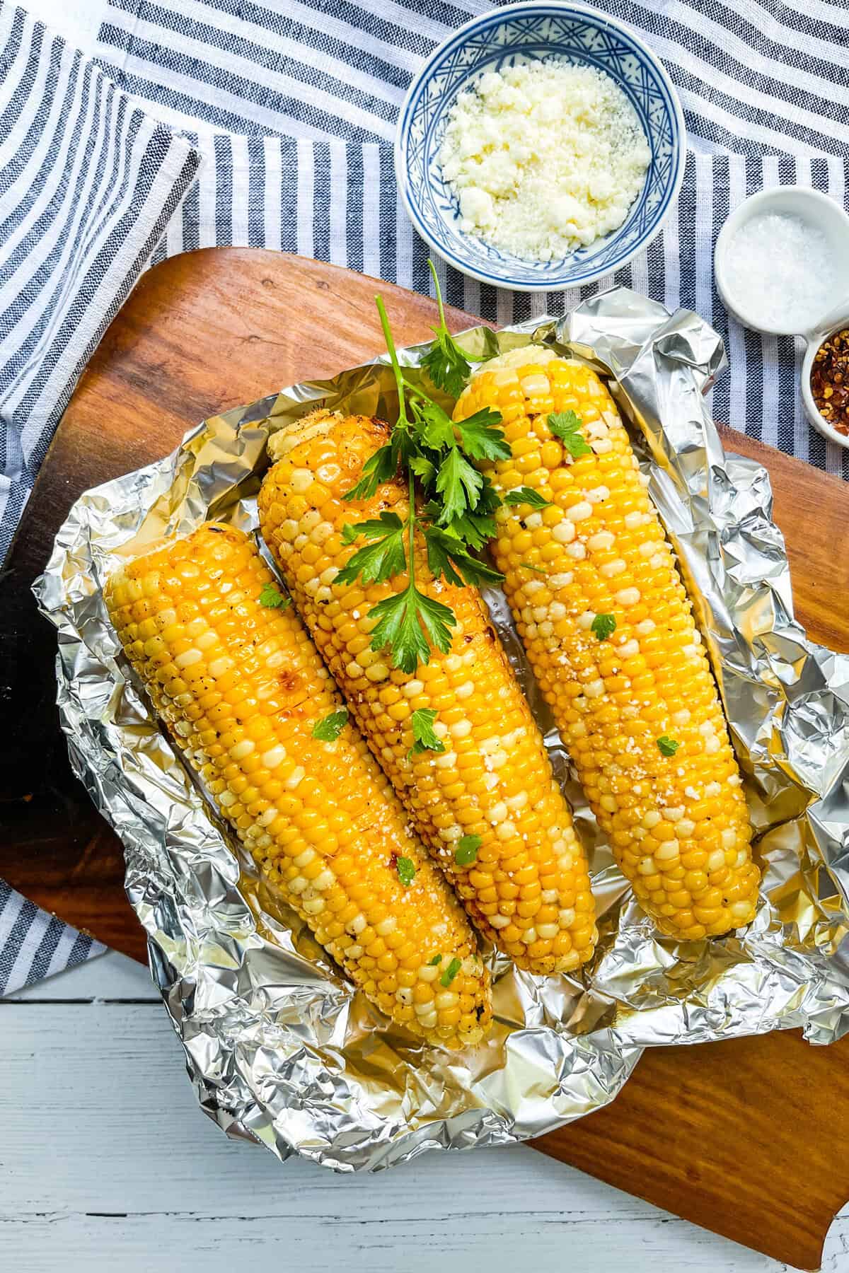 Three grilled corn on the cobs garnished with parsley rest on foil atop a wooden board, surrounded by bowls of crumbled cheese, salt, and red pepper flakes.