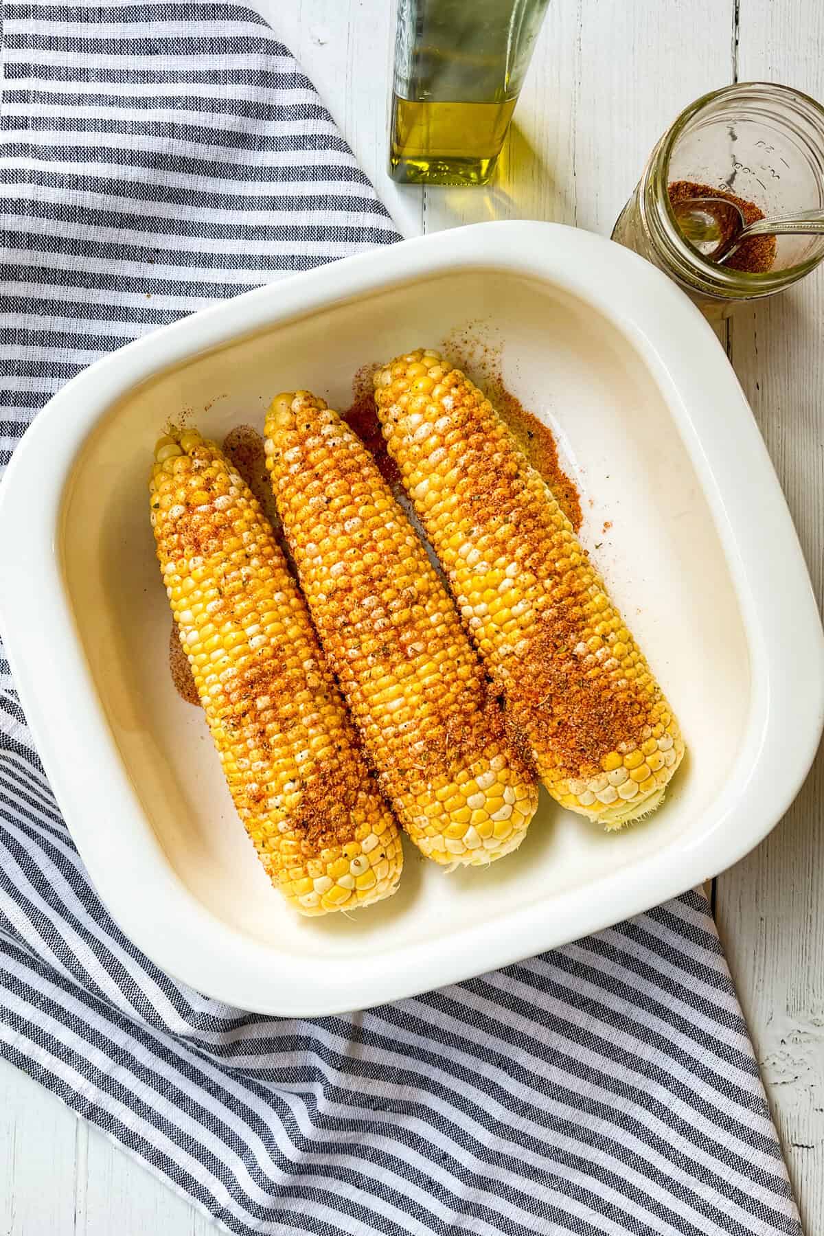 Three ears of grilled corn in a white dish are sprinkled with seasoning, placed on a striped cloth with a jar of spice and a bottle of oil nearby.