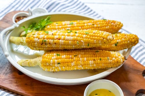 Four ears of grilled corn on the cob, garnished with chopped herbs, are arranged on a white plate, with a small bowl of melted butter nearby for dipping.