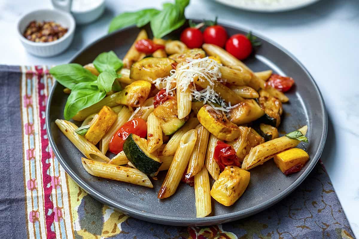 A serving a zucchini pasta on a dark plate with fresh cherry tomatoes, zucchini, squash, and fresh basil.