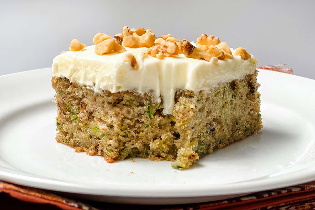 A slice of moist zucchini cake with cream cheese frosting and chopped walnuts on top sits on a white plate.