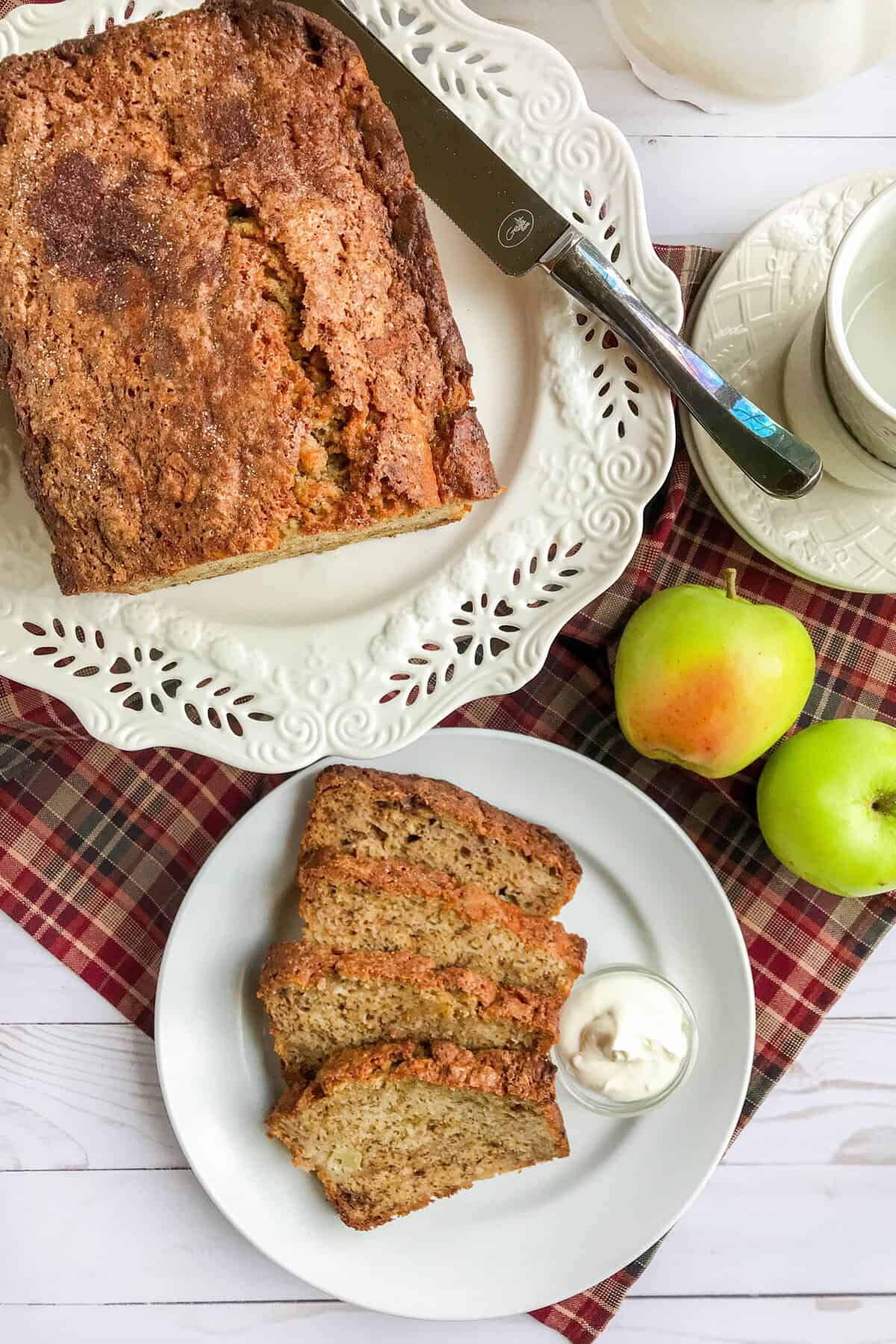 A loaf of apple banana bread on a decorative plate with a knife, three slices on a white plate with cream, two green apples, and a cup on a saucer atop a plaid cloth.