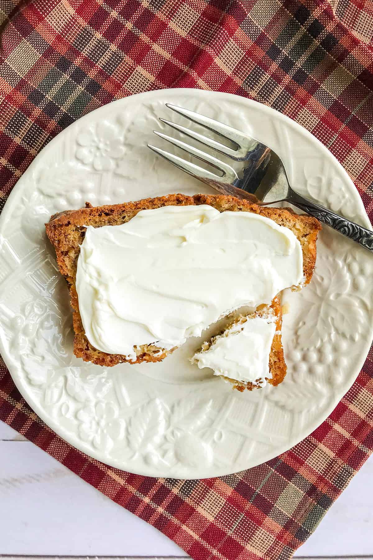 A slice of apple banana bread with cream cheese spread sits on a white plate, partially eaten, with a fork on the side and placed atop a red and brown plaid cloth.