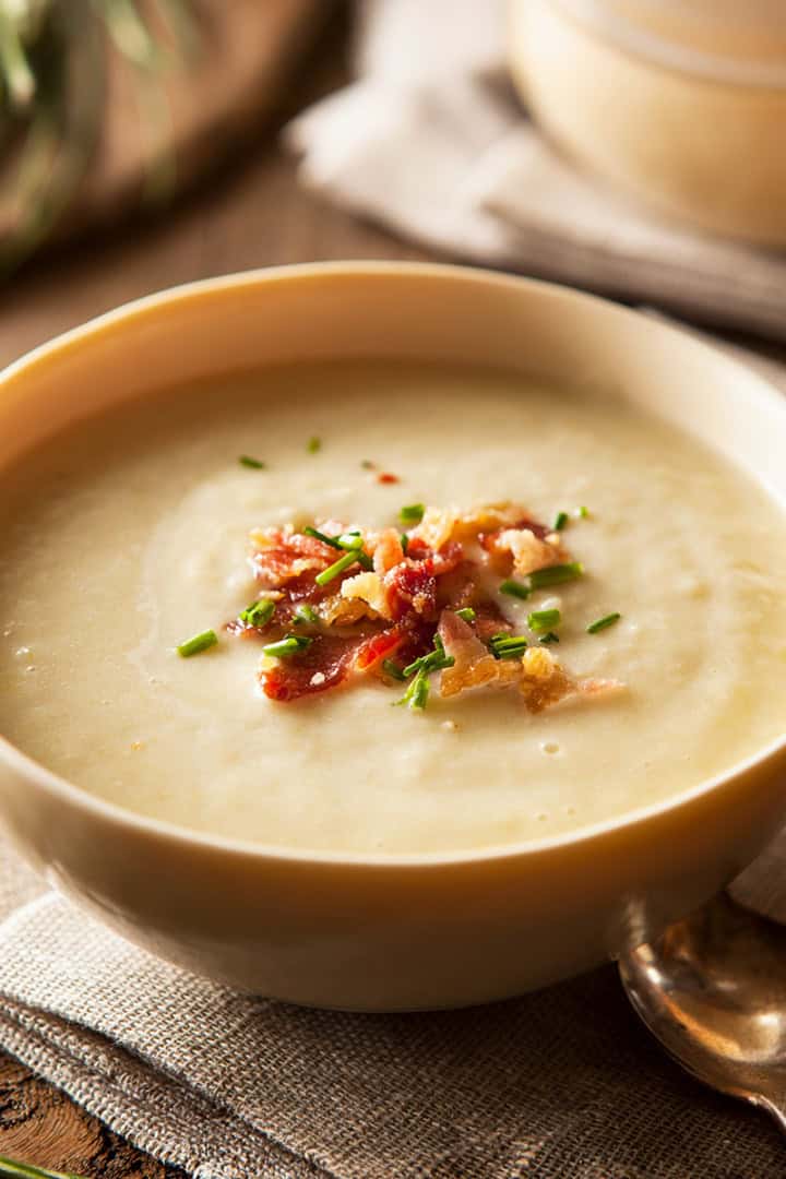 A bowl of creamy potato soup garnished with crispy bacon, chopped chives, and fried onions, placed on a napkin with a spoon beside it.