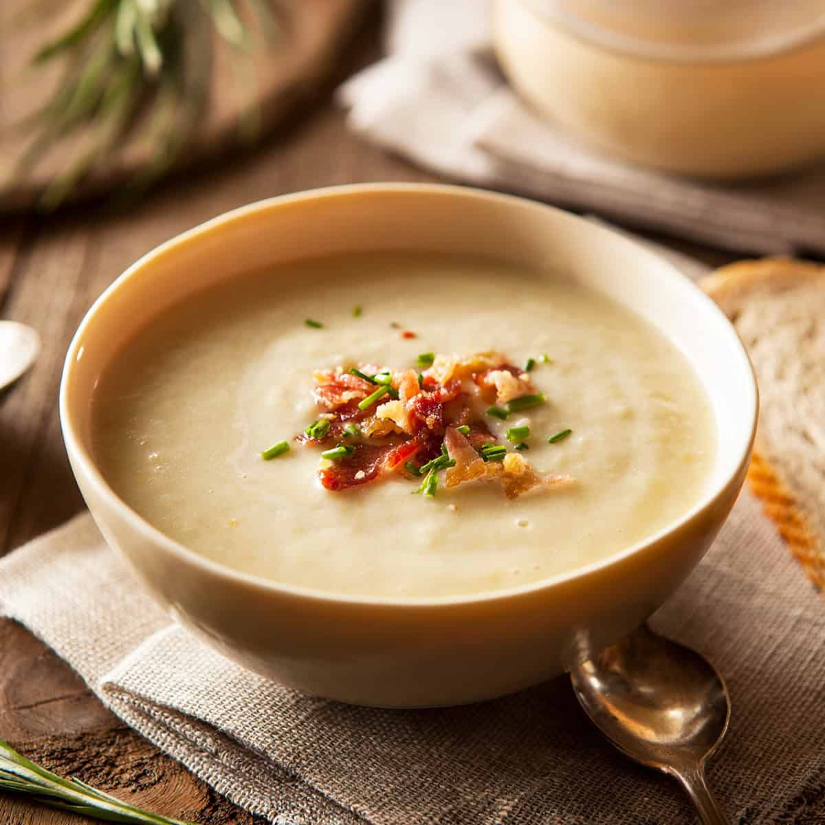 A bowl of creamy potato soup garnished with crispy bacon, chopped chives, and fried onions, placed on a napkin with a spoon beside it.