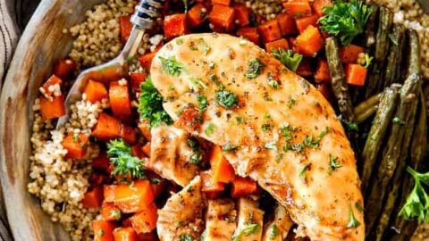 A bowl of cooked quinoa topped with sliced chicken breast, roasted carrots, and green beans—perfect for September sheet pan dinners—garnished with parsley. A fork rests in the bowl.