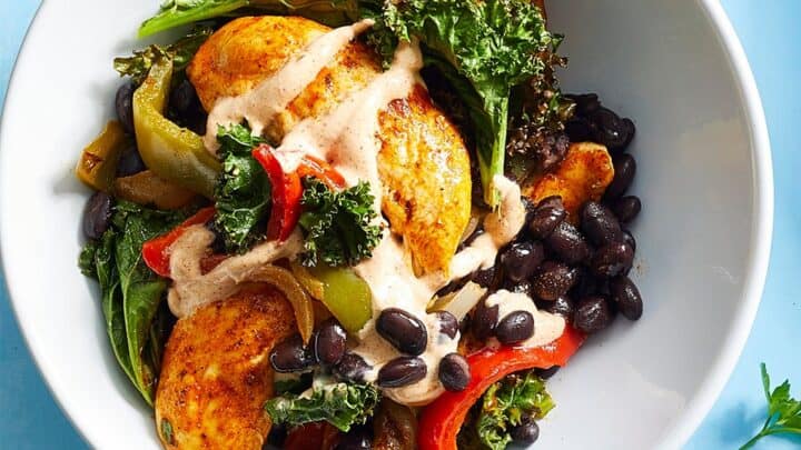 A white bowl filled with seasoned chicken, black beans, sautéed bell peppers, kale, and a creamy sauce on top—perfect inspiration for September sheet pan dinners—set on a light blue surface.