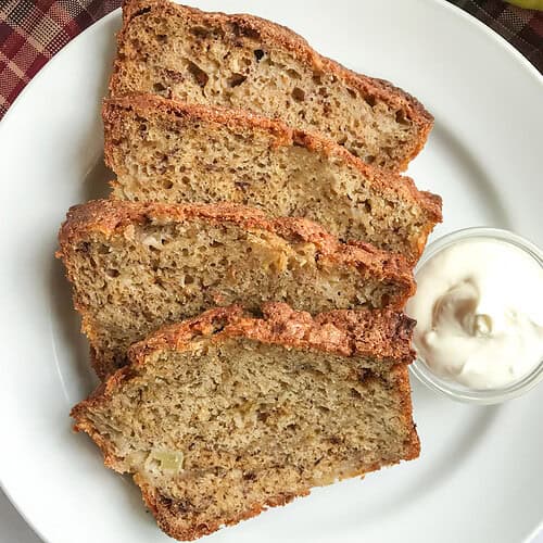 Four slices of apple banana bread are arranged on a white plate with a small glass bowl of cream or yogurt on the side, showcasing this delicious apple banana bread recipe.