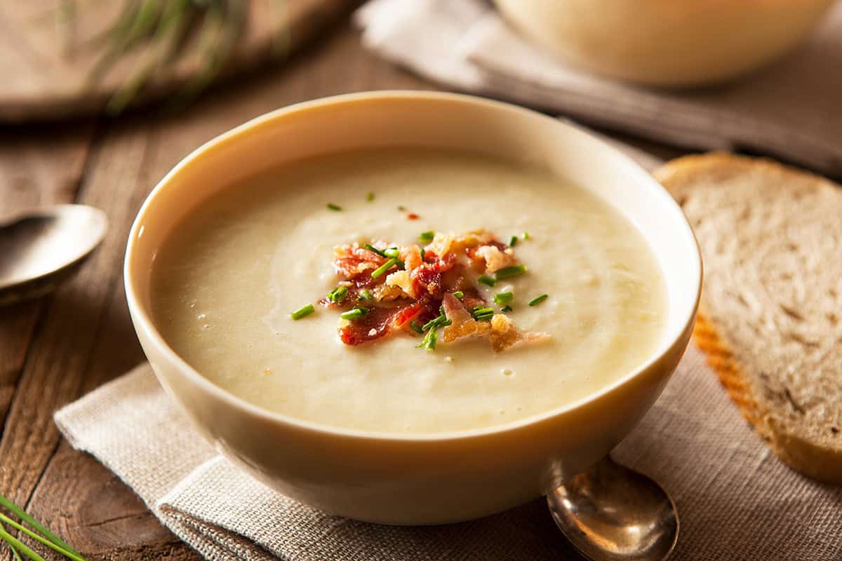 A bowl of creamy potato soup topped with bacon bits and chopped chives, served with a slice of bread and a spoon on a wooden table.