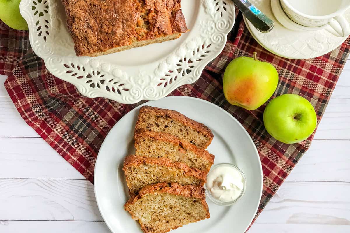 A plate with three slices of apple banana bread and a small cup of cream sits next to a loaf of apple banana bread, green apples, and a teacup on a red plaid cloth. Perfect for trying a new apple banana bread recipe!.