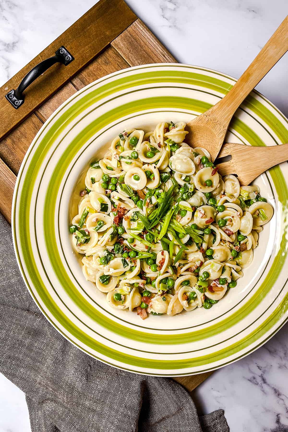 A plate of orecchiette pasta with peas, bacon, and green onions-an inviting option for easy pasta recipes-served with wooden utensils on a green-striped plate atop a wooden tray.