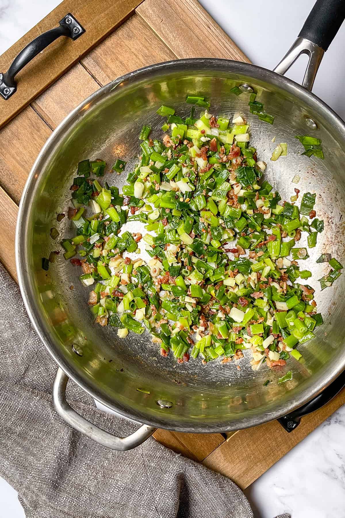 Chopped green onions and diced ingredients are being sautéed in a stainless steel pan on a wooden cutting board with a gray cloth nearby, perfect for easy pasta recipes or your next Weeknight Pasta creation.