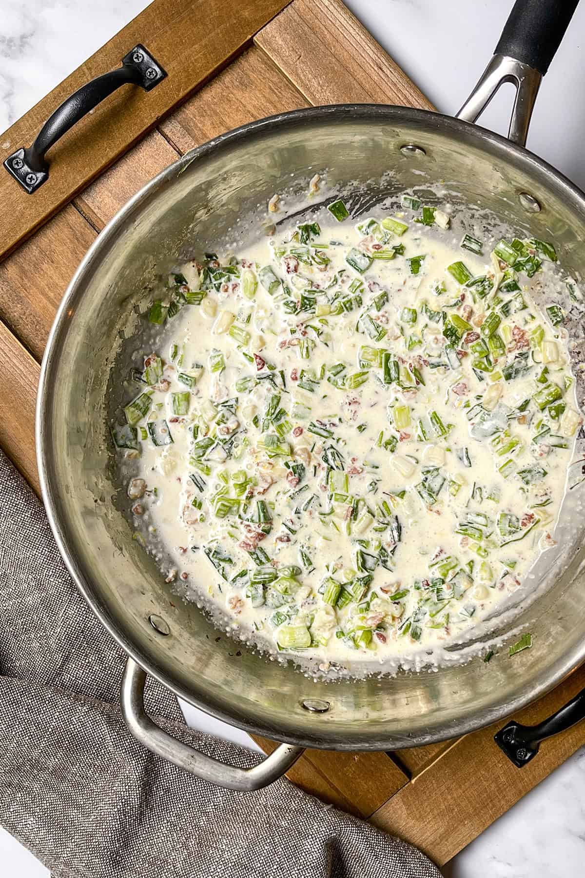 A stainless steel pan on a wooden tray contains a creamy mixture with chopped green onions and bacon pieces-perfect inspiration for your next Weeknight Pasta or pasta recipes.