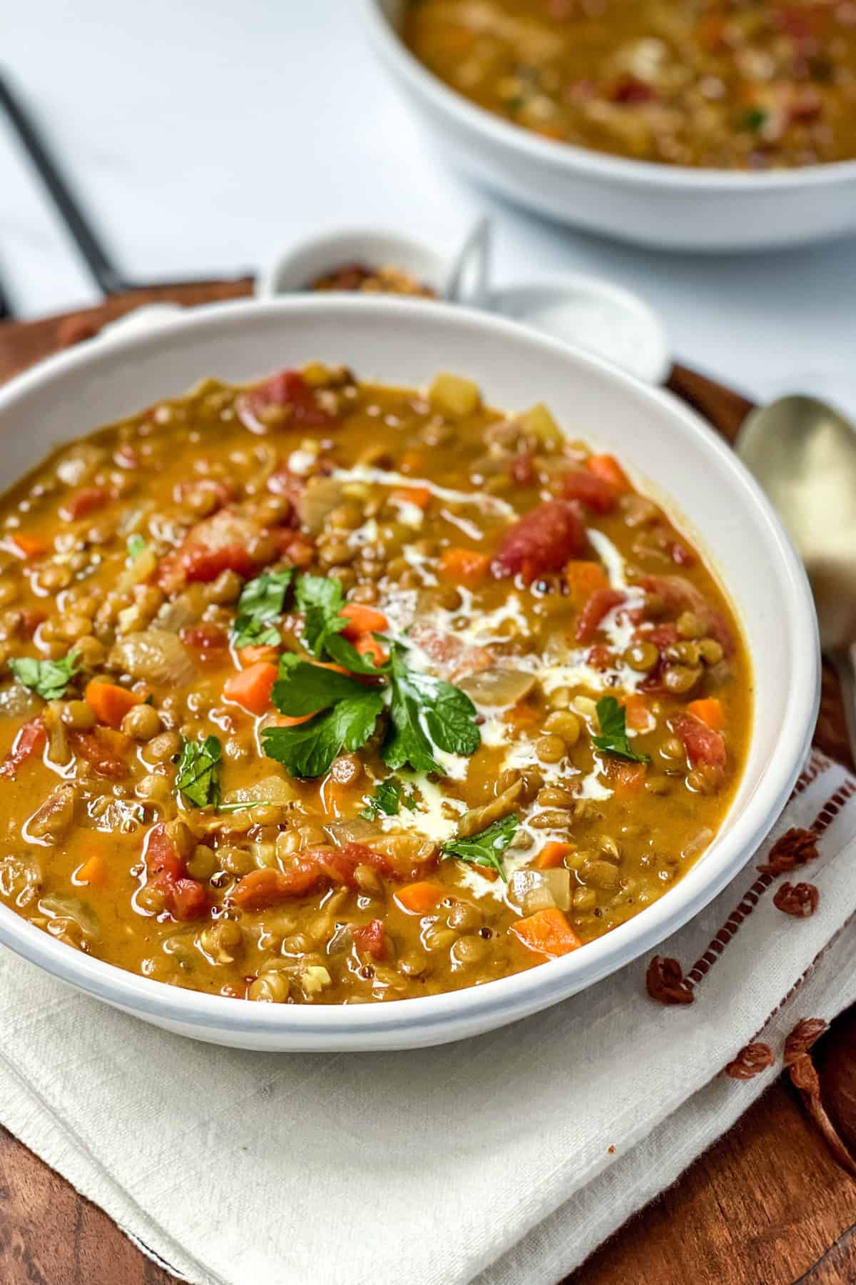 Serving of curry lentil soup in a white bowl garnished with fresh parsley and swirls of coconut milk with a spoon nearby.