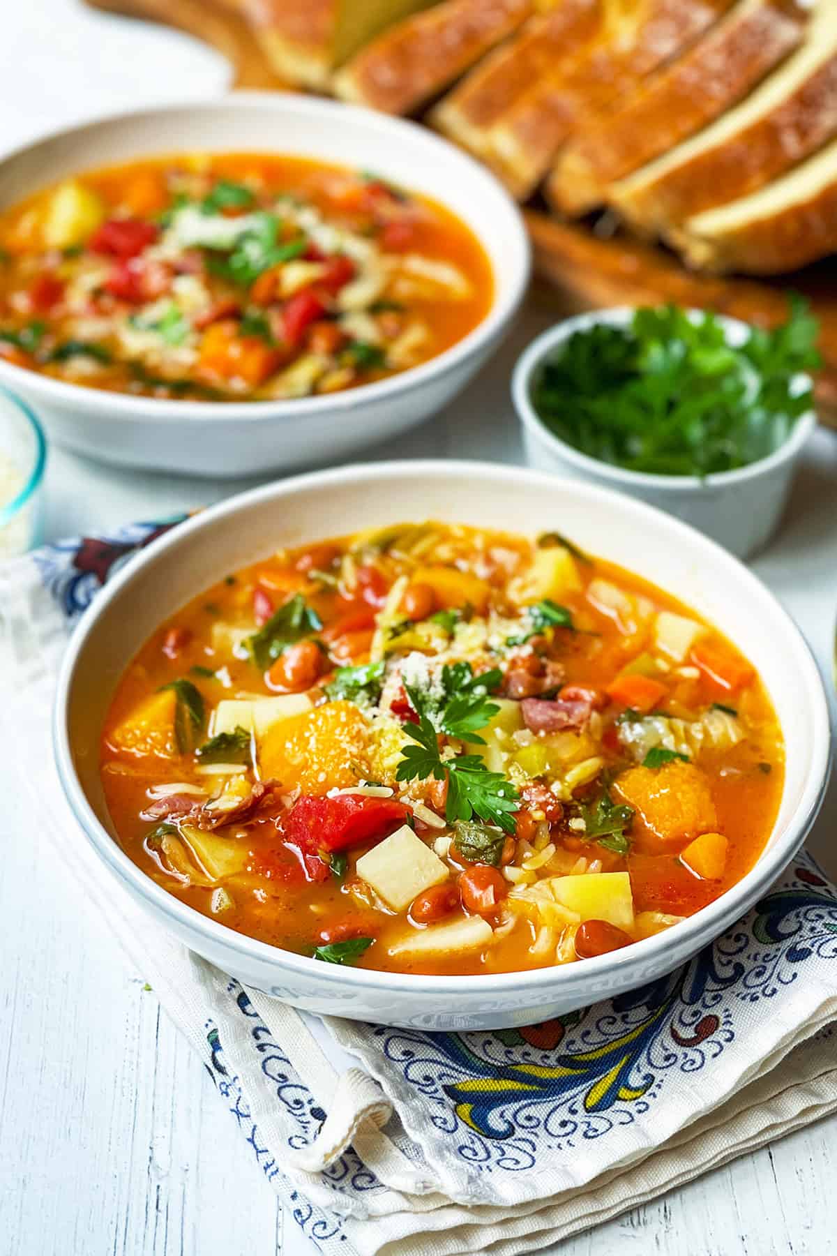 A bowl of minestrone soup recipe topped with herbs sits on a patterned napkin, with another bowl of soup, sliced bread, and a small bowl of parsley in the background.