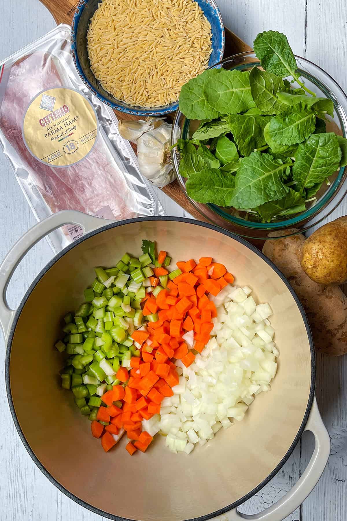 Chopped celery, carrots, and onion in a pot surrounded by orzo pasta, spinach, garlic, potatoes, and packaged pancetta on a white surface-perfect ingredients for a hearty minestrone soup recipe.