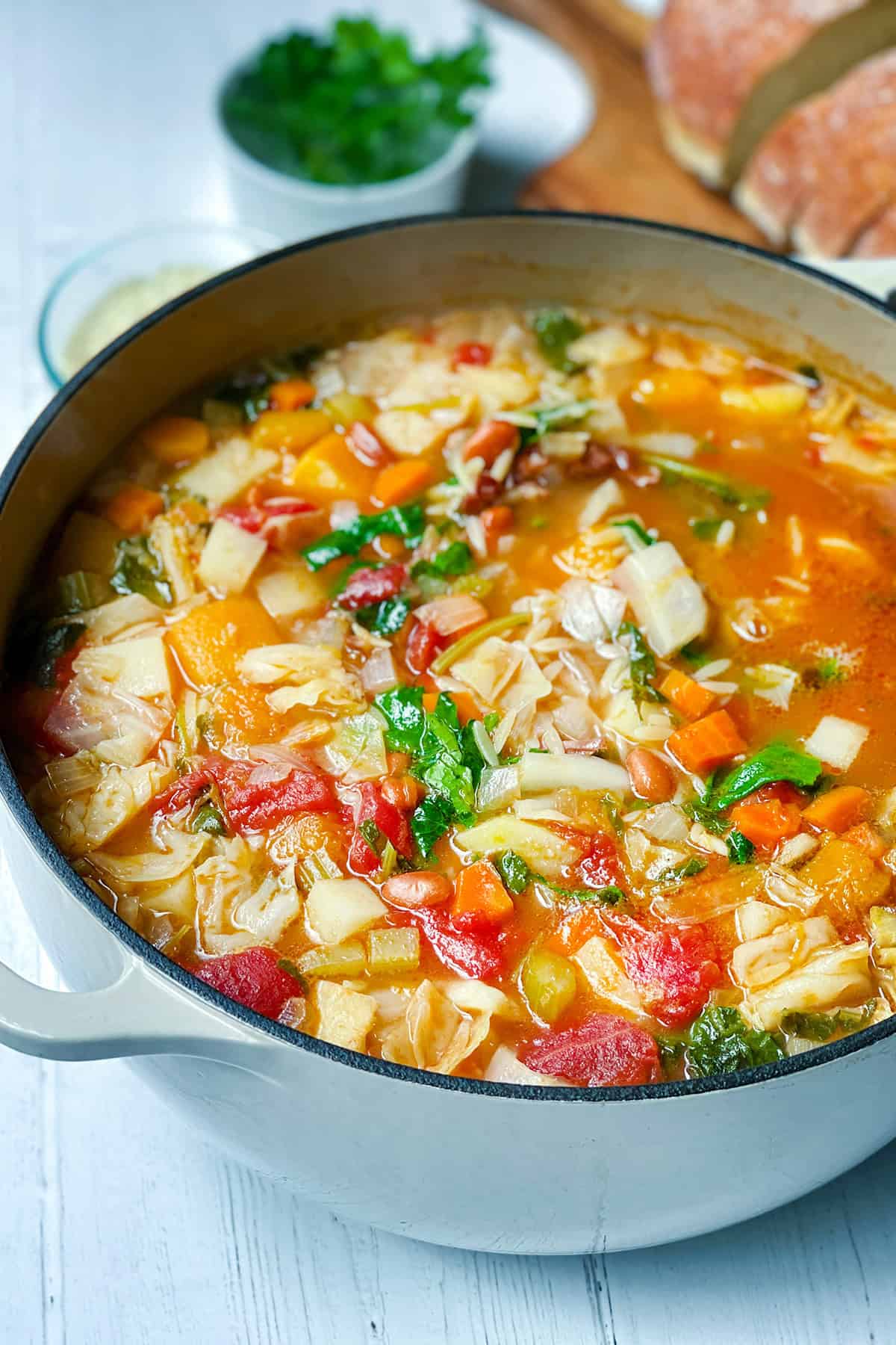 A large pot of minestrone soup recipe with cabbage, carrots, tomatoes, and greens sits on a white surface, with bread and herbs in the background.