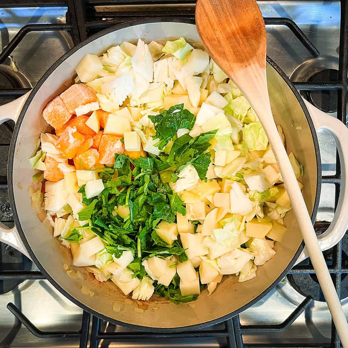 Chopped vegetables, including cabbage, carrots, potatoes, and greens, simmer in a pot on the stove with a wooden spoon resting on the side-perfect for starting your favorite minestrone soup recipe.
