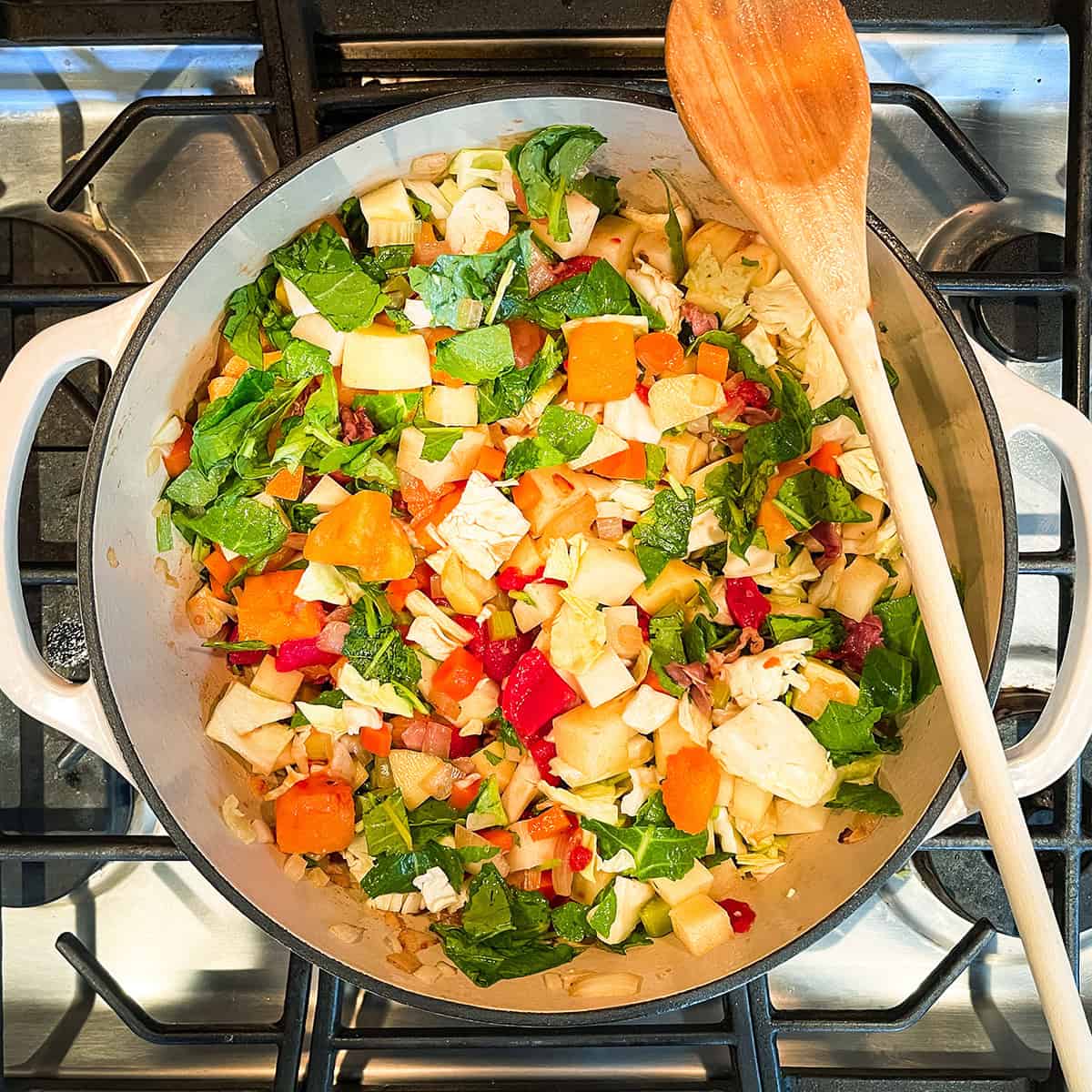A pot of chopped vegetables, including carrots, celery, potatoes, and greens, simmers on the stove with a wooden spoon resting on the side-the start of a classic minestrone soup recipe.