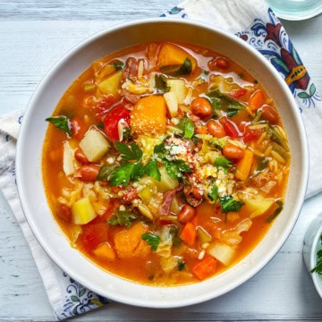 A bowl of classic minestrone soup recipe with beans, carrots, potatoes, tomatoes, greens, and grated cheese on top, placed on a patterned napkin.