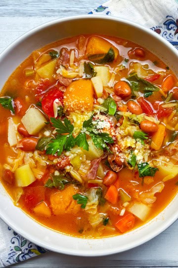 A bowl of classic minestrone soup recipe with beans, carrots, potatoes, tomatoes, greens, and grated cheese on top, placed on a patterned napkin.