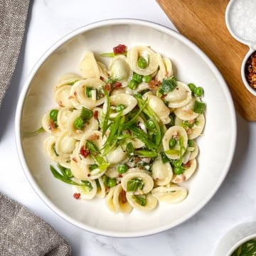 A white bowl of orecchiette pasta with green peas, chopped scallions, and bits of bacon or pancetta-an easy weeknight pasta recipe-sits on a white surface next to a grey napkin and a wooden board with seasonings.