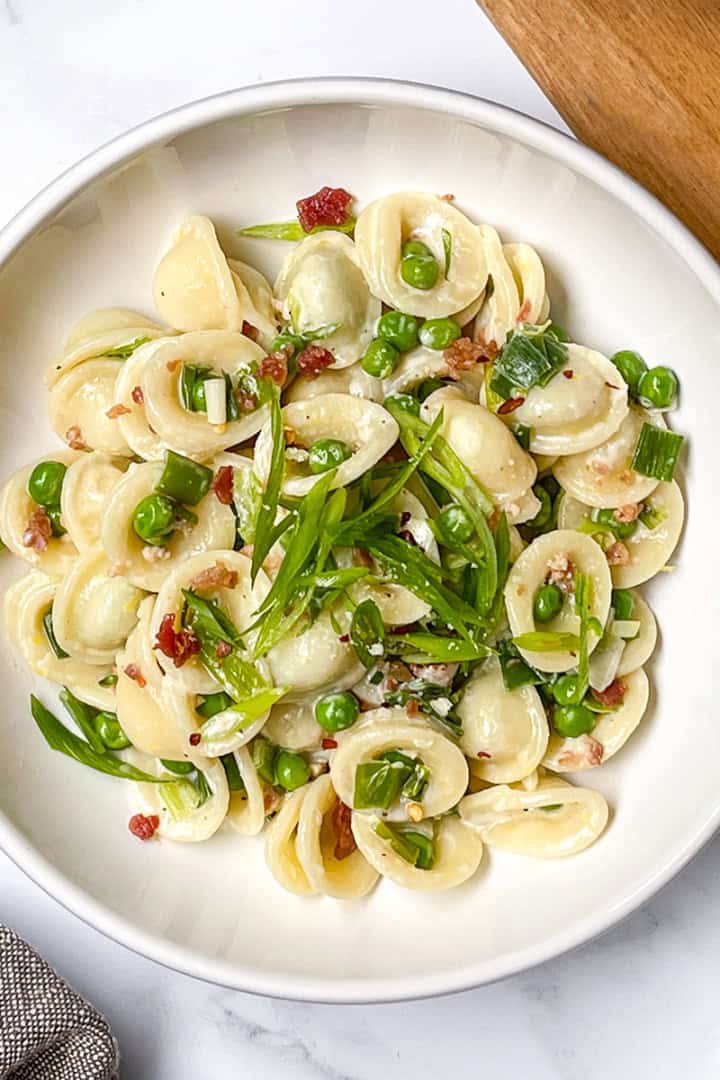 A white bowl of orecchiette pasta with green peas, chopped scallions, and bits of bacon or pancetta—an easy weeknight pasta recipe—sits on a white surface next to a grey napkin and a wooden board with seasonings.