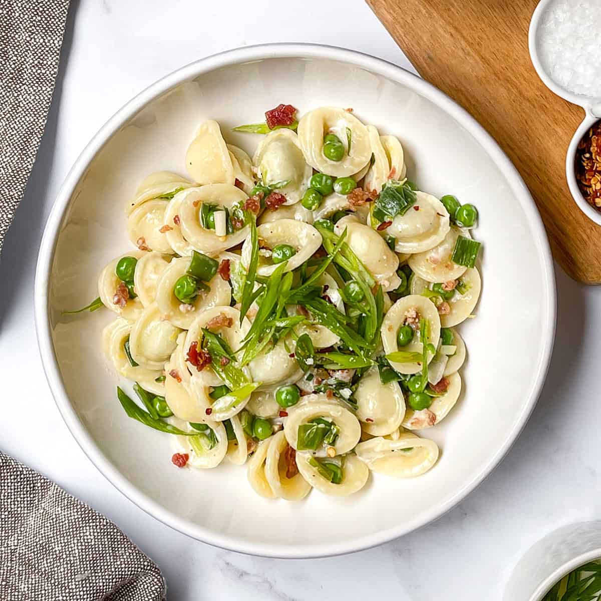 A white bowl of orecchiette pasta with green peas, chopped scallions, and bits of bacon or pancetta—an easy weeknight pasta recipe—sits on a white surface next to a grey napkin and a wooden board with seasonings.