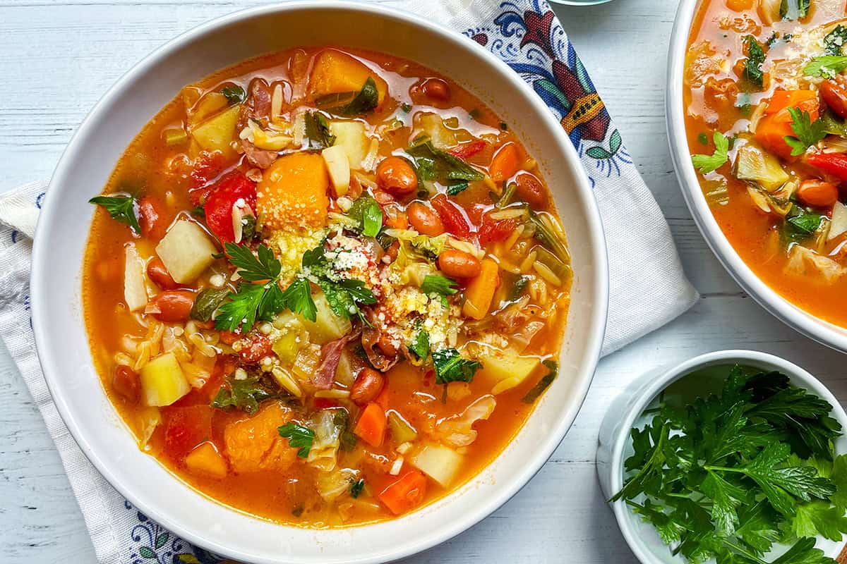 A bowl of hearty minestrone soup recipe with beans, potatoes, carrots, tomatoes, and greens, topped with grated cheese and fresh parsley, sits on a white table next to a small bowl of parsley.