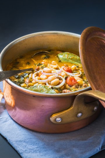 Copper pot filled with Ribollita, Tuscan Bean Soup, with crackers on the side, is resting on a linen ready for serving.