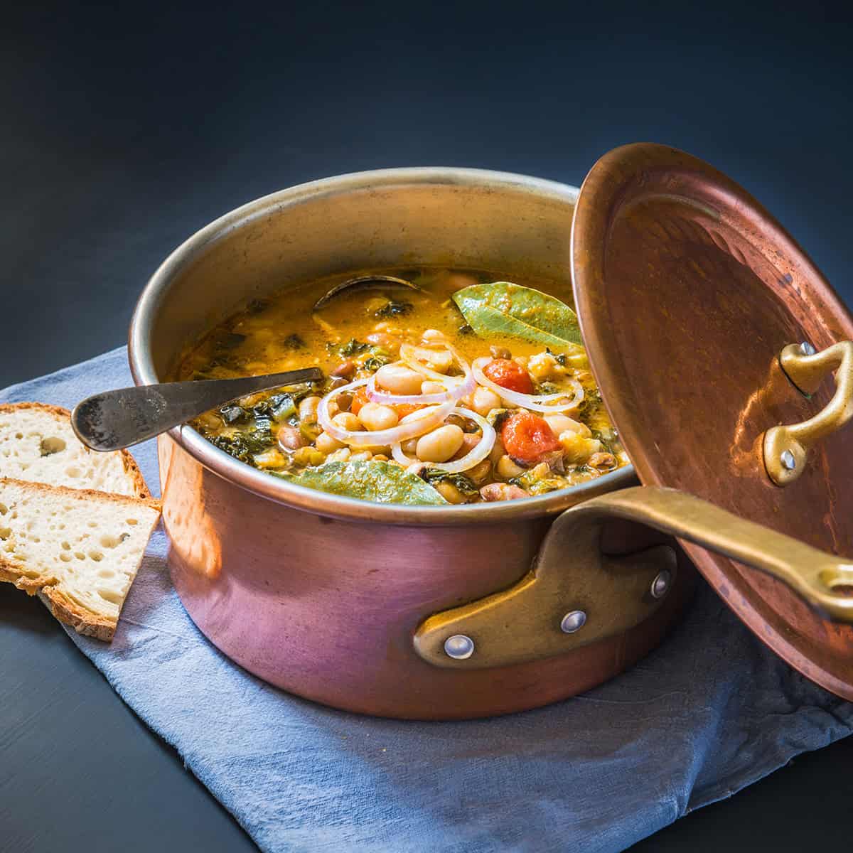 Copper pot filled with Ribollita, Tuscan Bean Soup, with crackers on the side, is resting on a linen ready for serving.