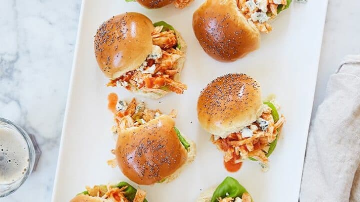 A rectangular white plate holds nine sliders with shredded chicken, lettuce, crumbled cheese, and poppy seed-topped buns&mdash;perfect for quick November 15 Minute Dinners. Some sliders have their tops slightly askew.
