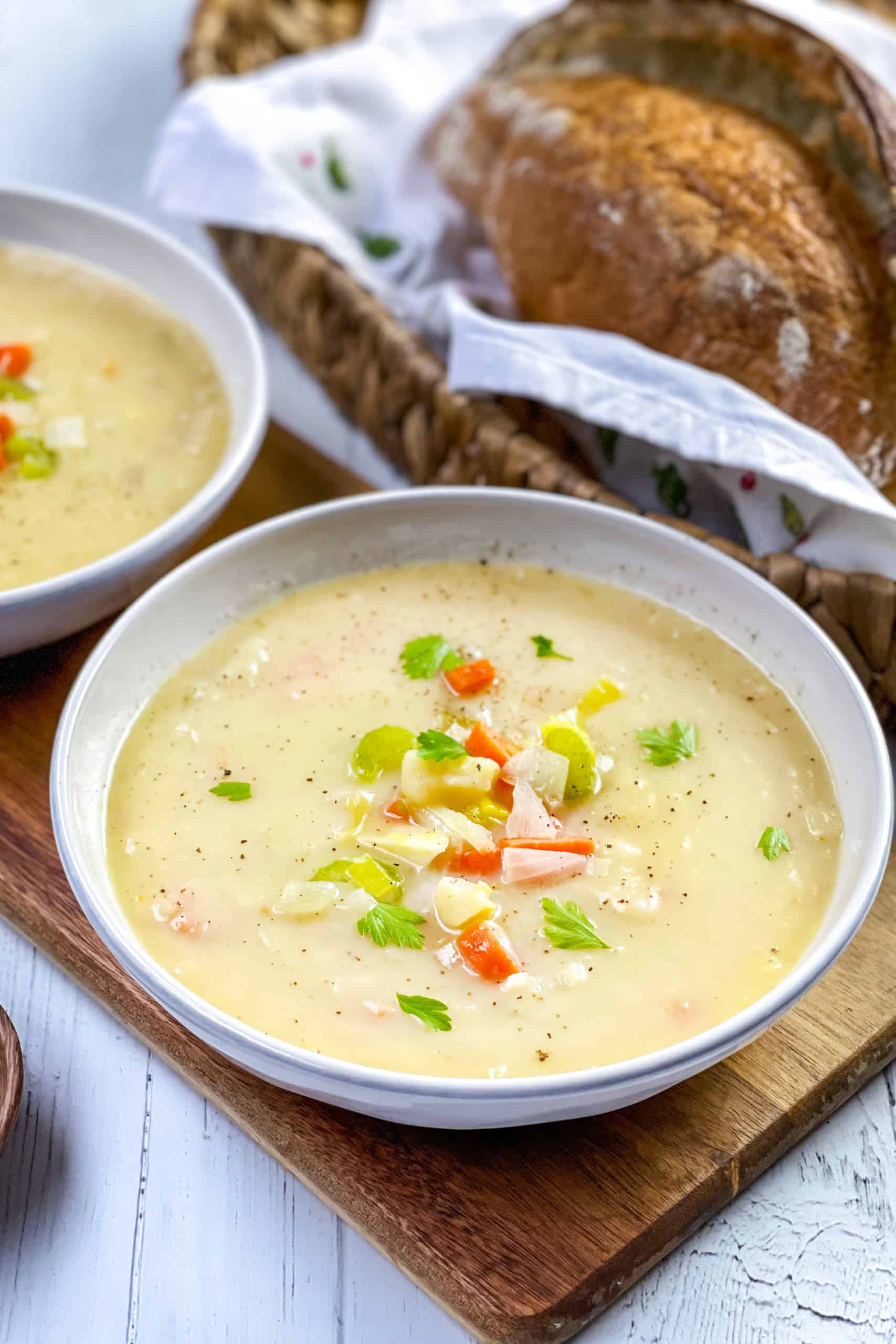 A bowl of creamy potato leek soup garnished with chopped celery, carrots, and herbs, placed next to a basket of sliced bread.