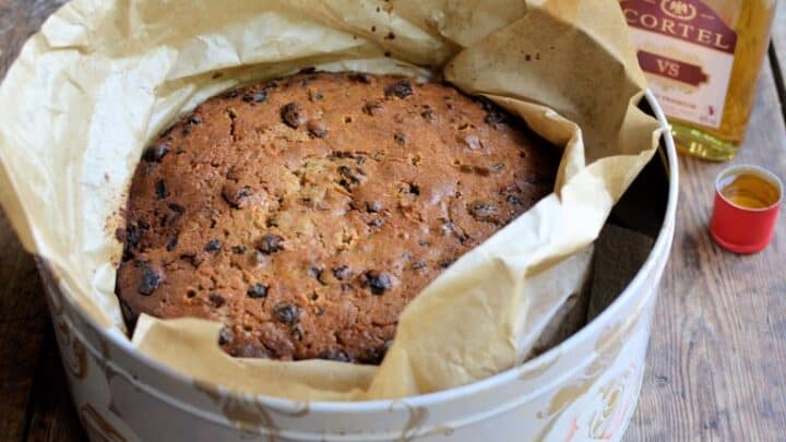 A round fruitcake in a lined cake tin sits on a wooden surface next to a bottle of brandy and a small filled cup, ready for Stir-Up Sunday tradition.