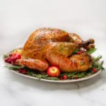 Top vertical view of roast turkey with apple cider on a serving platter garnished with fresh bay leaves, rosemary, whole apples, and cranberries.
