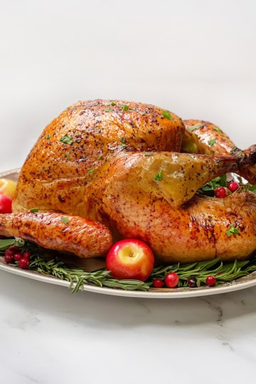 Top vertical view of roast turkey with apple cider on a serving platter garnished with fresh bay leaves, rosemary, whole apples, and cranberries.