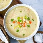 A bowl of creamy vegetable soup, inspired by classic potato leek soup, garnished with fresh parsley and filled with chunks of carrot, celery, and onion sits beside a bowl of salt on a wooden surface.