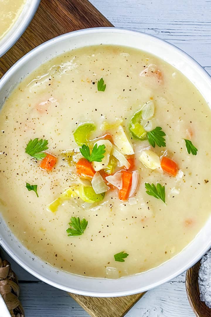 A bowl of creamy vegetable soup, inspired by classic potato leek soup, garnished with fresh parsley and filled with chunks of carrot, celery, and onion sits beside a bowl of salt on a wooden surface.
